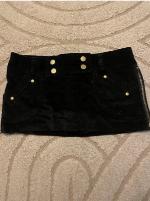 Svart kortkjol med guldknappar - Svart kort manchesterkjol med snygga guldknappar framtill och på fickorna. Kjolen har två framfickor och dragkedja i sidan. Perfekt för dig som gillar en trendig och cool stil. Har shorts under dragkedjorna på sidan kan öppnas🔥🔥 Riktig vintage😊 Använd gärna köp nu❤️