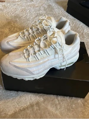 Nike Air Max 95 vita sneakers - Snygga Nike Air Max 95 sneakers i helvitt med mesh och mockadetaljer. Skorna har synliga Air-enheter i sulan och ikoniska vågiga paneler på sidan. Perfekta för dig som gillar klassisk streetwear och vill ha en clean look.