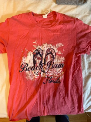 Beach Bum t-shirt Florida - Snygg t-shirt med tryck av flipflops och texten 'Beach Bum Florida' på framsidan. Skön passform och klassisk rund halsringning. Perfekt för dig som gillar avslappnad strandstil.