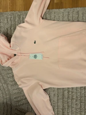 Ljusrosa hoodie från Lacoste - HELT OANVÄND, KÖPT ALLDELES NYSS! Anledningen till att jag säljer denna är för att jag redan har en och fick denna i present! Och vill få bort den så snabbt som möjligt. HELT ÄKTA! STORLEK M/4 