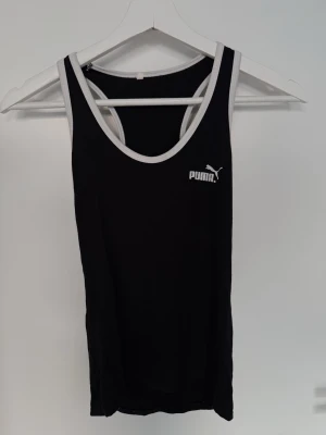 Linnee från Puma - Snygg svart linnetopp från Puma med vita kanter och klassisk logga på bröstet. Modellen är ärmlös och har en sportig look med rundad halsringning.  Använd några fåtal gånger och är i bra skick förutom att texten bak på ryggen blivit lite sliten (se bild).