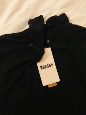 Svart jumpsuit från Bodssy - Snygg svart Semhar jumpsuit från Bodssy med hög krage. Knäppning med flera knappar vid nacken. Tight passform och stretchigt material(Modal & elastane) som sitter otroligt skönt på kroppen. Perfekt för dig som vill ha en stilren och trendig look. Storlek XS.