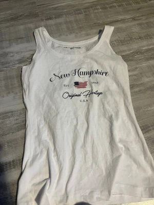 Vit linnetopp från Reaction S - Snygg vit linnetopp från Reaction med tryck framtill som säger 'New Hampshire' och har en amerikansk flagga. Toppen är ärmlös och har rund halsringning. Perfekt till sommaren och enkel att matcha med jeans eller shorts.