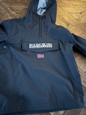 Svart vindjacka från Napapijri - Svart vindjacka från Napapijri med huva och halv dragkedja framtill. Stor ficka på bröstet med Napapijri-logga och norsk flagga. Jackan har en sportig look och är tillverkad i slitstarkt material, perfekt för blåsiga dagar. Har ett jätte litet hål i fickan vid bröstet