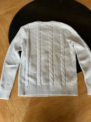 Ljusgrå kabelstickad tröja Hollister - Ljusgrå kabelstickad tröja från Hollister med rund halsringning och ribbade muddar. Tröjan har ett diskret Hollister-logotyp broderat på bröstet och är gjord i mjukt material. Klassisk kabelstickning framtill ger en snygg och tidlös look.