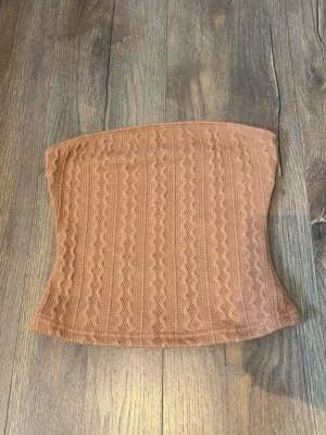 Brun stickad tubtopp - Säljer en snygg brun stickad tubtopp med kabelstickat mönster. Toppen är åtsittande och har en mjuk, stretchig känsla som gör den bekväm att bära. Perfekt att styla med jeans eller kjol för en trendig look.