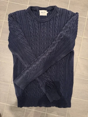 Mörkblå kabelstickad tröja NN07 - Klassisk kabelstickad tröja från kvalitetsmärket NN07 (No Nationality 07).  En tidlös tröja i en snyggt tvättad marinblå (washed navy). Modellen heter "Cable" och är tillverkad i kraftig, högkvalitativ bomull som gör att den andas bra och fungerar lika bra över en skjorta som till ett par jeans.  Väl omhändertagen och har kvar sin passform.  Nypris: ca 1 400 kr.