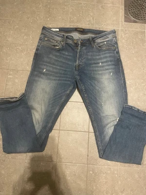 Blå slitna jeans från Jack & Jones - Säljer ett par blå jeans från Jack & Jones med snyggt slitna detaljer och ljusare partier framtill. Klassisk femficksmodell med normal midja och raka ben. Jeansen har en patch med logga bak i midjan och är tillverkade i bomull med lite stretch.