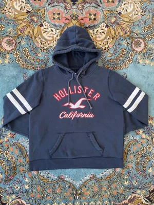 Mörkblå Hollister hoodie - Mörkblå hoodie från Hollister med vit och röd text på bröstet där det står 'Hollister California' och en måslogga. Har huva med dragsko, känguruficka och vita dubbla ränder på ärmarna. Perfekt för en avslappnad och sportig stil. Passar S-M, vid frågor eller funderingar skriv gärna!