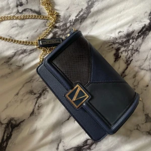  Victoria's Secret - Snygg mörkblå axelväska från Victoria's Secret med guldfärgad kedja och elegant V-detalj framtill. Väskan är i skinnimitation med inslag av strukturmönster och har ett rymligt fack med dragkedja och kortfack inuti. Perfekt för att lyfta din outfit. Aldrig använd