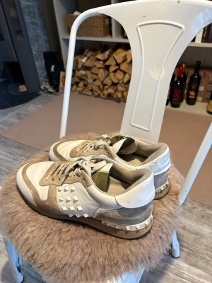 Valentino Garavani Rockstud sneakers - Snygga sneakers från Valentino Garavani med beige och vita paneler i mocka och läder. Ikoniska nitar längs sidan och hälen ger en edgy vibe. Skorna har klassisk låg siluett och cool kontrast mellan material och detaljer. De är litet hål på insidan av hälen,därav lågt pris! 