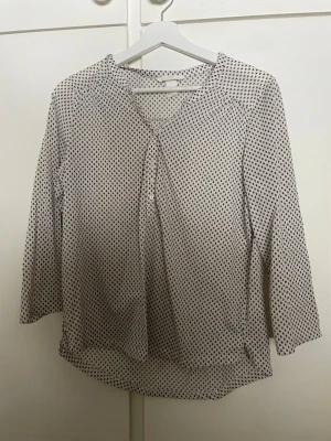 Vit blus med svart mönster från H&M - Vit blus från H&M med små svarta bladliknande mönster. Blusen har v-ringning, knappdetalj framtill och trekvartsärm. Tillverkad i ett lätt och luftigt material som ger en avslappnad och snygg look.