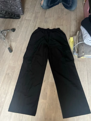 Svarta cargobyxor med vida ben - Säljer ett par svarta cargobyxor med bred passform och stora fickor på sidorna. Byxorna har elastisk midja med dragsko och är tillverkade i ett mjukt tyg som känns bekvämt. Perfekta för en avslappnad och trendig look.