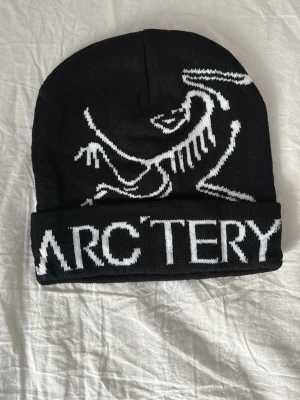 Svart mössa från Arc'teryx - Säljer en svart mössa från Arc'teryx med vit logga och text framtill. Mössan är ribbstickad och har uppvikt kant. Materialet är mjukt och stretchigt, perfekt för kalla dagar. Snygg och enkel design som passar till det mesta.