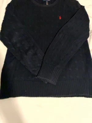 Mörkblå kabelstickad tröja Polo Ralph Lauren - Kabelstickad mörkblå tröja från Polo Ralph Lauren med rund halsringning och lång ärm. Klassisk röd logga broderad på bröstet. Tröjan har ribbade muddar vid ärmslut och nederkant. Perfekt för dig som gillar tidlös och stilren design.