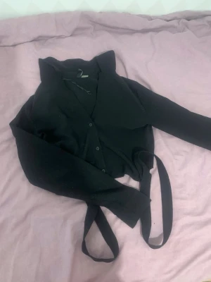 Svart omlottkofta med knytband - Snygg svart omlottkofta med långa ärmar och knytband i midjan. Koftan har en klassisk v-ringning och knappar framtill. Perfekt att styla över ett linne eller t-shirt för en trendig lager-på-lager-look.