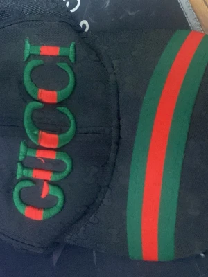 Svart Gucci keps med logga - Säljer en svart keps från Gucci med tydlig grön och röd logga broderad framtill samt klassiska gröna och röda ränder på skärmen. Kepsen har diskret GG-mönster i tyget och är tillverkad i ett slitstarkt material. Perfekt för dig som vill ha en stilren och lyxig accessoar.