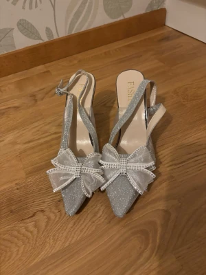 Glittriga silverpumps med rosett - HELT NYA! Aldrig använda. Snygga klackskor i silver med glittrigt material, spetsig tå och öppen häl. Dekorerade med  rosetter framtill och små strasstenar. Mellan stor/Smal klack och justerbar rem runt hälen. Perfekta för bal, bröllop förlovningen eller annan festlighet 