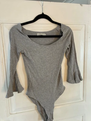 Grå ribbad body från Abercrombie - Säljer en grå ribbad body från Abercrombie & Fitch med långa ärmar och trendiga volangdetaljer vid ärmsluten. Bodyn har en bred, rundad urringning och knäppning i grenen. Perfekt för dig som gillar en enkel men snygg stil.