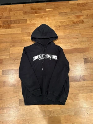 Svart True Religion hoodie - Säljer en svart hoodie från True Religion med stor vit logga på bröstet och en fet vit grafisk detalj på ryggen. Klassisk huva med dragsko och ribbade muddar. Perfekt för dig som gillar streetwear och vill sticka ut lite extra.