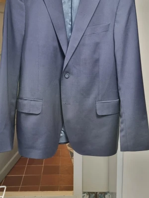 Mörkblå kostym från Oscar Jacobson - Stilren mörkblå kostym från Oscar Jacobson med klassisk passform, dubbelknäppt med två knappar och tre fickor framtill. Fodrad insida med diskret logotyp. Perfekt för en dressad look och passar till skjorta eller t-shirt.