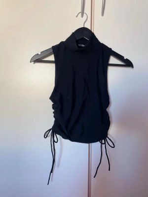 Svart haulterneck topp med snörning i sidan - Snygg haulterneck topp med trendig snörning i sidorna, perfekt att styla med höga jeans eller kjol. Materialet känns mjukt och stretchigt, vilket gör den bekväm att bära hela dagen. Det står att det är en storlek L men den känns som en storlek S.