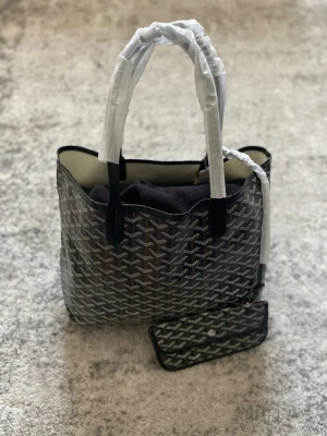 Svart shoppingväska med mönster - Stilren väska med rutigt mönster och en medföljande plånbok. Aldrig använd och i mycket fint skick, dustbag medföljer även. Hör gärna av dig vid frågor!