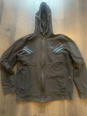 Svart hoodie från BOSS med dragkedja - Snygg svart hoodie från BOSS med hel dragkedja och huva. Har coola turkosa och vita detaljer på bröstet och axlarna. Två fickor framtill och diskret BOSS-logga på vänster sida. Perfekt för en chill och stilren look. Storlek M men passar nog S