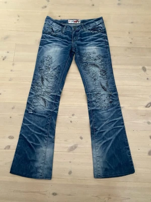 Blå bootcut jeans med broderi - Säljer ett par blå bootcut jeans från Nudie Jeans med snygga broderade detaljer och slitningar på framsidan. Jeansen har låg midja, klassisk femficksmodell och är tillverkade i denim. Perfekta för dig som vill sticka ut med coola detaljer.