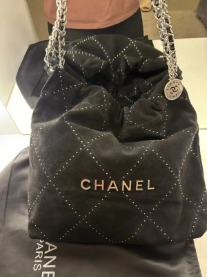 Svart handväska  - Snygg svart handväska från Chanel i mjukt mockaliknande material med silverfärgade kedjehandtag. Väskan har ett rutmönster med små nitar och en stor CHANEL-logga framtill. Medföljer original dustbag och en rund silvercharm med logotyp.