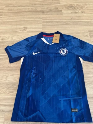 Chelsea FC 25/26 - Chelsea FC matchtröja i blått från Nike med klubbmärke på bröstet och vit Nike-logga. Tröjan har vita detaljer vid kragen och sidorna samt diskreta mönster i tyget. Perfekt för fotbollsträning eller att bära på läktaren.
