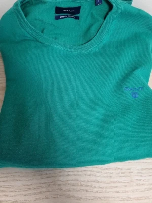 Grön tröja från GANT - Snygg grön tröja från GANT med rund halsringning och diskret broderad logotyp på bröstet. Tröjan är tillverkad i stretchig bomull och har en klassisk passform som är enkel att matcha med jeans eller chinos.