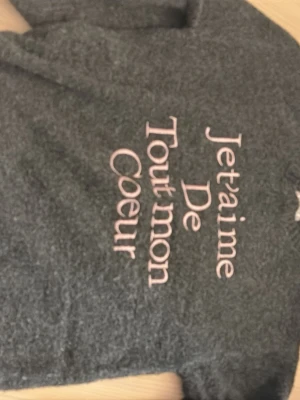 Mörkgrå stickad tröja Gina Tricot - Använt en gång,Tröjan är en mysig mörkgrå stickad tröja från Gina Tricot med rosa texten 'Je t'aime de tout mon coeur' på framsidan. Tröjan har rund halsringning och långa ärmar. Perfekt för dig som gillar trendiga och bekväma plagg med ett gulligt budskap.