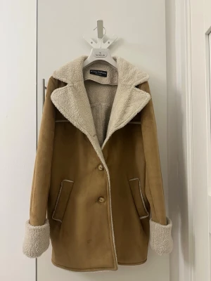 Camel shearling rock från Daisy Fashions - Snygg camelbrun rock från Daisy Fashions med mjukt, vitt teddyfoder. Rocken har bred krage, uppvikta ärmslut och två fickor framtill. Stängs med två knappar och har en avslappnad, rak passform. Perfekt för kalla dagar och ger en trendig look.