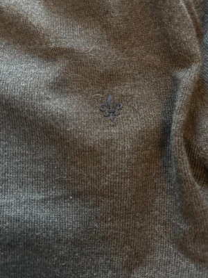 Brun stickad tröja från Morris - Säljer en stilren grå stickad tröja från Morris i 100% merinoull. Tröjan har rund halsringning och en liten broderad lilja på bröstet. 