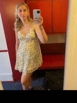 Blommig kortklänning - Söt kortklänning från Na-kd med smala axelband och v-ringning. Klänningen är vit med små blommor i beige och grönt, har en luftig passform och är perfekt för utekväll eller vardagsklänning