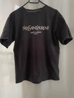 Blyertsgrå Yves Saint Laurent t-shirt - Snygg vintage  blyerts grå t-shirt från Yves Saint Laurent med klassisk logga tryckt i vitt på bröstet. T-shirten har rund halsringning och korta ärmar. Perfekt för dig som gillar stilrena och ikoniska plagg. Inget märke i nacken. Tjockt tyg 