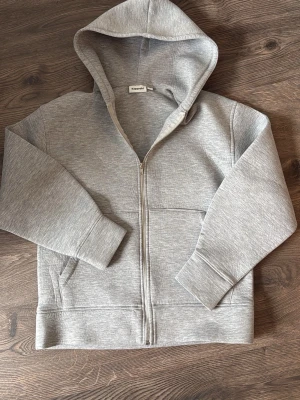 Grå zip hoodie från Kappahl - En stilren grå hoodie med dragkedja från Kappahl. Hoodien har en klassisk huva, två fickor framtill och breda muddar vid ärmslut och nederkant. Tillverkad i mjukt sweatshirtmaterial som är perfekt för en avslappnad look. Oversized passform.