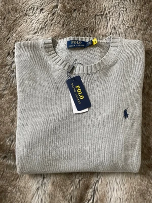 Ljusgrå stickad tröja Polo Ralph Lauren - Snygg ljusgrå stickad tröja från Polo Ralph Lauren med rund halsringning och klassisk blå logga broderad på bröstet. Tröjan har ribbade muddar vid hals, ärmslut och nederkant. Perfekt för dig som gillar stilrena och tidlösa plagg. Nypris ca 2000, mitt pris 699