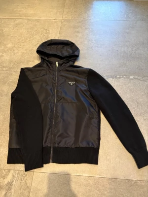 Svart hoodie från Prada med dragkedja - Säljer en svart hoodie från Prada med dragkedja och huva. Kroppen är i glansigt material medan ärmar och muddar är stickade. Prada-loggan sitter på bröstet. Perfekt för dig som vill ha en snygg och exklusiv hoodie med coola detaljer.