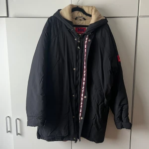 Svart parkajacka från Helly Hansen - Säljer en svart vinterjacka från Helly Hansen med beige foder och huva. Jackan har knappar och dragkedja framtill samt en dekorativ röd och vit rem. Perfekt för kalla dagar och har en avslappnad, loose passform. Mörkblå nästintill svart