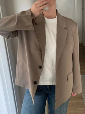 Beige oversized kavaj från Gina Tricot - Stilren beige kavaj från Gina Tricot i oversized modell. Klassisk design med två knappar framtill, breda slag och två fickor med lock. Tillverkad i ett mjukt tyg som ger en avslappnad men ändå dressad look. Perfekt för att lyfta vilken outfit som helst. Färgen Taupe (mellan brun och beige). Storlek 38, oversized. Med prislapp.
