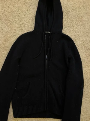 Marinblå stickad hoodie Brandy Melville - Marinblå stickad hoodie från Brandy Melville med dragkedja framtill och två fickor. Tröjan har ribbade muddar vid ärmslut och nederkant samt justerbar dragsko i huvan. Enkel och stilren design som funkar till det mesta.