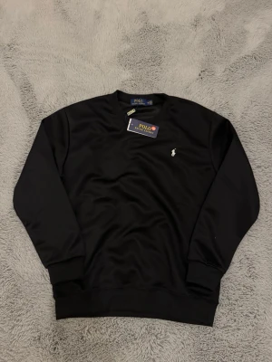 Svart sweatshirt från Polo Ralph Lauren - Stilren svart sweatshirt från Polo Ralph Lauren med klassisk rund halsringning och broderad vit logga på bröstet. Tröjan har ribbade muddar vid ärmslut och nederkant. Perfekt för en avslappnad och trendig look. Det står storlek M men den är liten i storlek så skulle nog säga den passar bättre på någon som bär Xs/S (160-170cm)