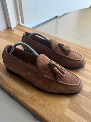 Bruna mocka loafers - Snygga bruna loafers i mocka med dekorativa tofsar framtill och klassisk rundad tå. Skorna har en låg svart sula och stilren design som passar perfekt till jeans eller chinos. Perfekt för dig som vill ha en tidlös och trendig look.