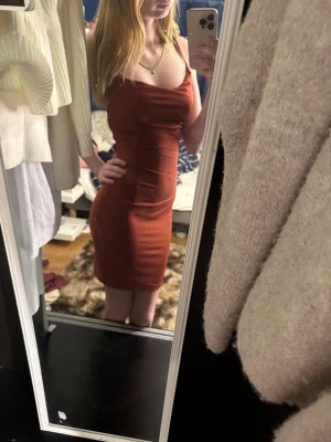 Rostbrun tight klänning med smala axelband - Säljer en snygg rostbrun klänning med smala axelband och v-ringad hals. Klänningen är kort och har en figurnära passform som framhäver siluetten. Perfekt för dig som gillar minimalistisk stil och vill ha något enkelt men stilrent.