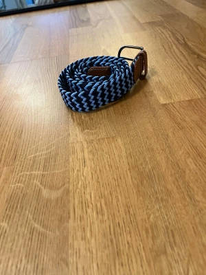 Flätat blått och svart från onewear - Snyggt flätat bälte i blått och svart med detaljer i brunt läder och silverspänne. Perfekt accessoar för att lyfta din outfit med en sportig och modern touch. Bältet är elastiskt och passar de flesta midjor.