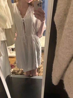 Vit kortklänning med snörning - Säljer en vit kortklänning med snörning framtill och v-ringad hals.aldrig använt då jag glömde den som jag skulle ha med mej till min konfirmation. Klänningen har en avslappnad passform och är tillverkad i ett lätt material, perfekt för varma dagar. Ärmarna är korta och klänningen har en enkel, stilren design.