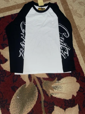 Corteiz longsleeve - Säljer en svartvit långärmad t-shirt med coolt grafiskt tryck längs båda ärmarna. Klassisk raglanmodell med svart krage och vita paneler framtill och baktill. Perfekt för dig som gillar streetwear och vill sticka ut lite extra.