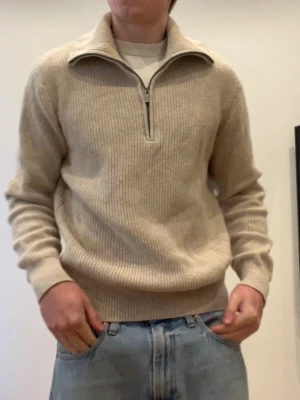 Beige stickad tröja med halvzip - Säljer en beige stickad tröja med halvzip och hög krage. Tröjan har ribbstickad struktur, långa ärmar och breda muddar vid ärmslut och nederkant. Perfekt för lager-på-lager och enkel att matcha med jeans.